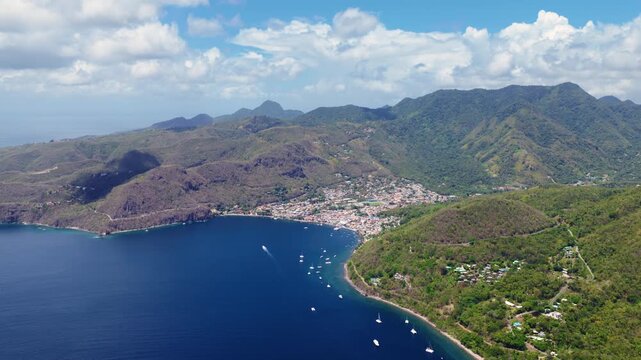 Stunning aerial view of Piton mountains rising above a coastal town in Saint Lucia. Yachts anchor in deep blue bay while lush green volcanic peaks stretch toward cloudy skies over the Caribbean.