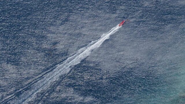 Aerial footage captures a red speedboat cutting through deep blue ocean waters leaving a dramatic white wake trail behind. The boat moves diagonally across calm sea surface.