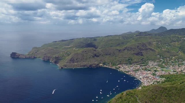 Stunning aerial view of iconic Piton mountains rising above lush green hillsides in Saint Lucia. Boats anchor in calm bay near colorful coastal town with dramatic volcanic cliffs meeting Caribbean.
