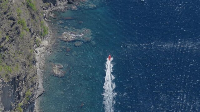 Aerial view of red speedboat creating white wake trail as it speeds along the dramatic Piton coastline in Saint Lucia. Crystal clear turquoise waters reveal rocky seabed near rugged volcanic cliffs.