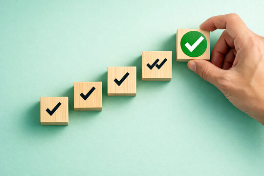 Wooden blocks showcasing checkmarks and a green symbol represent a step-by-step progression as a hand positions the final piece to finalize the project goal in a work space. Business growth success