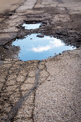 A large, water-filled pothole on a cracked asphalt road reflects a bright blue sky and white clouds, emphasizing road deterioration. © Jennifer
