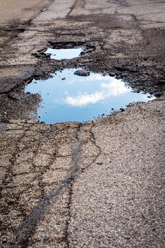 A large, water-filled pothole on a cracked asphalt road reflects a bright blue sky and white clouds, emphasizing road deterioration.