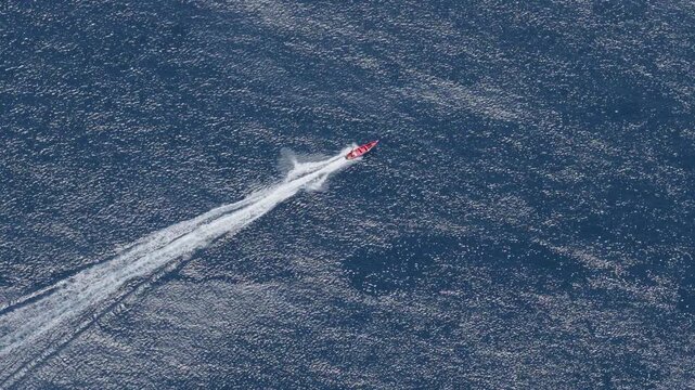 Aerial view of a red speedboat speeding across dark blue ocean water creating a long white wake trail. The boat moves diagonally through the sparkling sunlit sea surface.