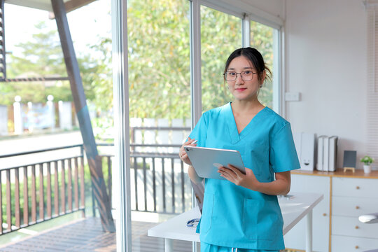 Young female medical professional wearing scrubs, holding a stylus with a tablet, looking at camera in a hospital setting