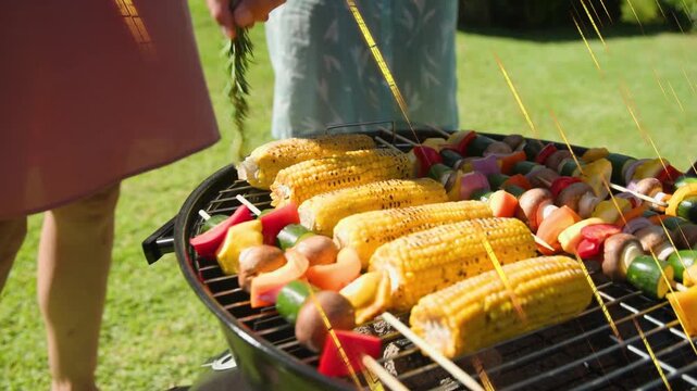 Man in gray shirt moving bowl, sprinkling marinade, brushing herb over corn, skewers for grilling