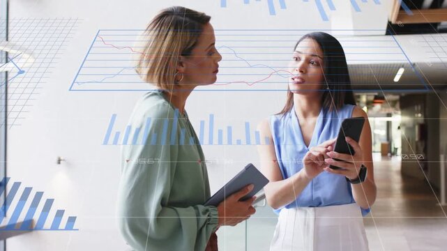 Two businesswomen standing in corridor, right woman tapping phone, showing data charts for review