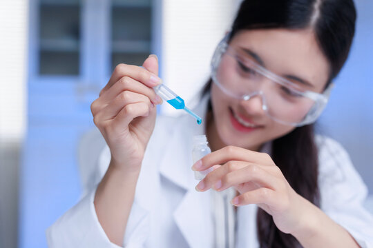 Young Asian scientist in lab coat and safety glasses dropping blue liquid from a pipette into a small vial, conducting research
