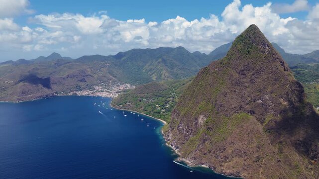 Stunning aerial view of the iconic Piton volcanic peak rising above the deep blue Caribbean Sea in Saint Lucia. Yachts anchor in the bay near Soufriere town with lush green mountains in background.