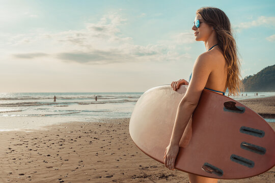 Female surfer with sunglasses holding surfboard on sunny beach looking at sea