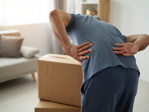 Person Bent Over Holding Lower Back in Pain While Picking Up Box in Modern Apartment