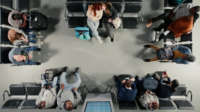 view of tired passengers sleeping in airport next to departure board showing delayed flights. Multiethnic people napping on chairs in lounge next to FIDS timetable screen, high angle shot
