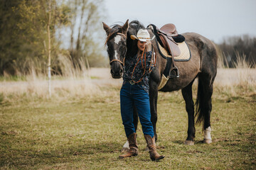 Female equestrian wearing cowboy hat and boots stands beside brown horse in grassy field with trees and cloudy sky in the background © Emvats