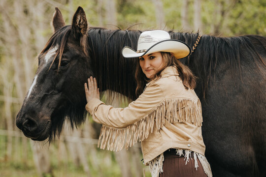 Young woman in fringed leather jacket and cowboy hat gently touches the side of a large black horse in a natural outdoor setting with greenery in the background