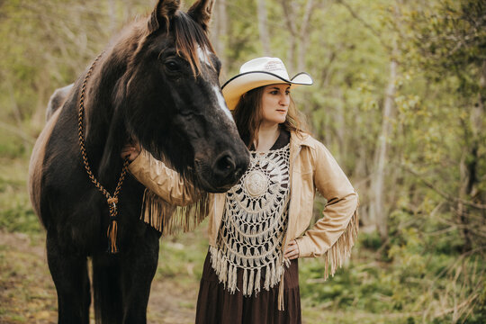 Young woman in cowboy hat and fringed jacket stands beside a black horse in a lush green forest, showcasing a rustic outdoor setting with natural light