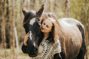 Fototapeta premium Young woman with long brown hair gently embraces a black horse in a forested area, sunlight filtering through the trees creating a serene atmosphere