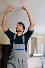 Professional handyman in overalls inspects residential ceiling for repairs and maintenance, looking...