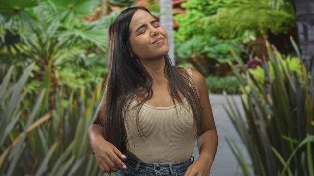 Woman face squinting, eyes closed and head tilted while adjusting waistband of jeans on building courtyard walkway lined with plants; discomfort.