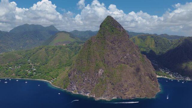 Stunning aerial footage of the iconic Piton volcanic peaks in Saint Lucia with lush green tropical rainforest slopes meeting deep blue Caribbean waters. Boats dot the coastline near scenic bays.
