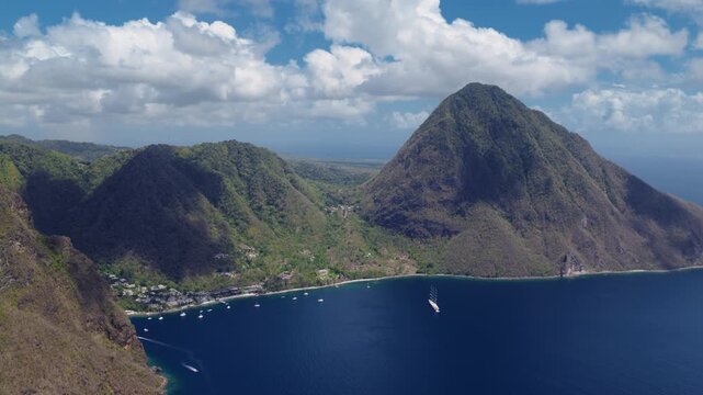Aerial drone footage of iconic Piton mountains rising above tranquil Caribbean bay in Saint Lucia. Lush green volcanic peaks tower over deep blue waters with sailboats anchored near tropical.