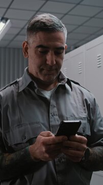 Officer With Arm Tattoos Checks Notifications and Social Media on Smartphone by Locker, While Other Staff Members Undress After Airport Shift. Workplace Communication and Relaxation. Vertical Shot.