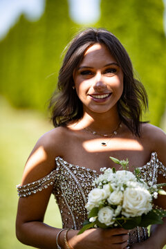 Young graduate wearing a dress made out of small gems, holding a flower and lit by the sun picking between a shade making pattern of shaded and sunny areas