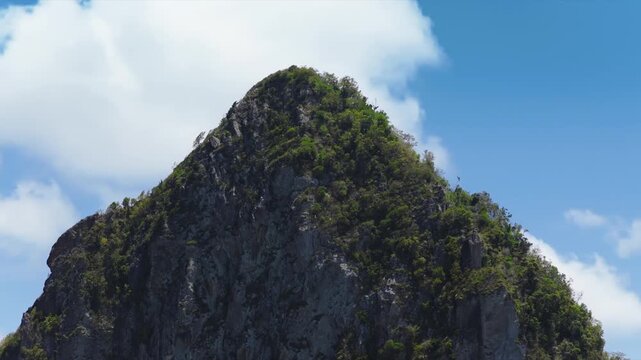 Dramatic volcanic Piton peak covered in lush green tropical vegetation rises majestically against bright blue sky with white clouds drifting past in Saint Lucia Caribbean island paradise.