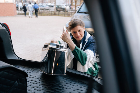 Young man in retro jacket puts shiny metal jerrycan into car trunk. View from inside vehicle showing preparation for road trip or fuel emergency. Urban parking lot background with natural light.