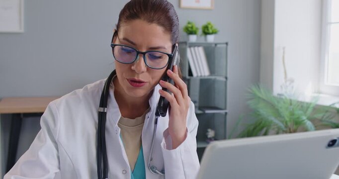 Experienced woman doctor giving medical consultation via mobile phone or advice for treatment, posing at workplace in medical clinic, busy chatting with patient. Health center assistant