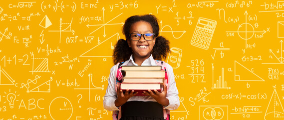 A young student stands happily with books in a school environment. Various math and science symbols fill the background. The girl wears glasses and has curly hair tied back. © Prostock-studio
