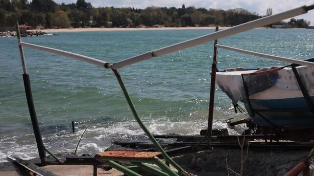 old abandoned boat with metal frame covered in tarp on rocky shore with turquoise sea waves sandy beach and green trees in background
