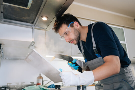 Professional worker wearing safety goggles and gloves cleaning kitchen extractor hood filter using steam cleaner, ensuring sanitation and hygiene in commercial kitchen environment.