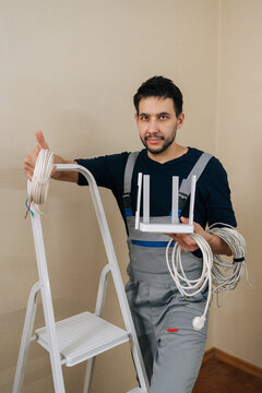 Service worker in overalls holding modern wireless router and network cables, giving thumbs up sign while standing next to ladder, ready to set up internet.