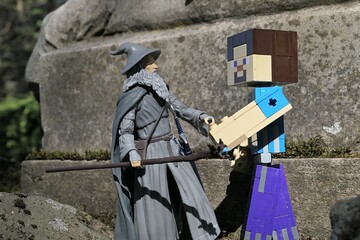 Obraz premium Minecraft action figure of main character Steve shaking hands with figure of wizard Gandalf from Lord Of The Rings and Hobit books by J.R.R. Tolkien in front of stone pedestal, spring sunshine. 