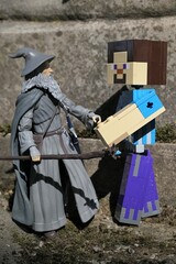 Obraz premium Minecraft action figure of main character Steve shaking hand with action figure of wizard Gandalf from Lord Of The Rings and Hobit books by J.R.R. Tolkien in front of stone pedestal, spring sunshine. 