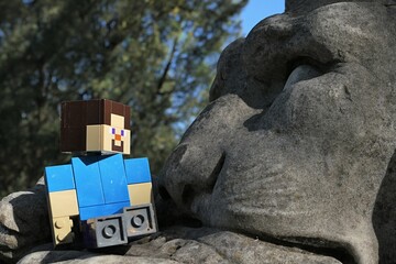 Obraz premium Action figure of main Minecraft Character Steve looking on a face of a giant lion statue in park, spring blue skies in background. 