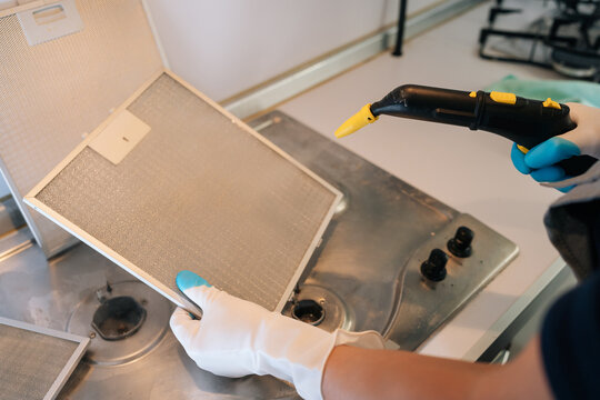 Person wearing protective gloves cleaning dirty metal grease filter from cooker hood with steam cleaner, ensuring kitchen hygiene and removing accumulated fat and grime from kitchen appliances.