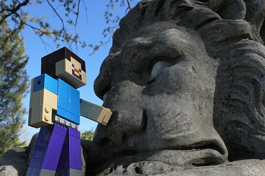 Action figure of main Minecraft Character Steve touching a nose of a giant lion statue in park, spring blue skies in background.