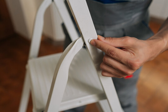 Close-up view of man hand opening white metal stepladder. Worker in grey overalls preparing equipment for home improvement or repair. Shallow depth of field focuses on hand and hinge.