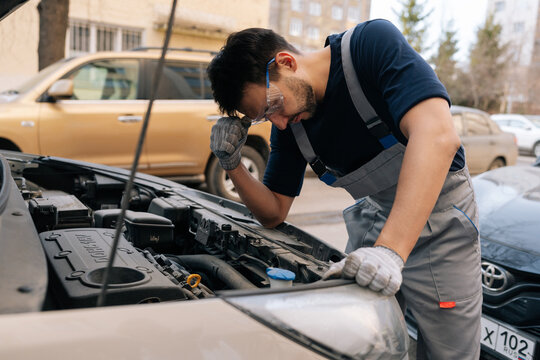 Mechanic troubleshooting complex car engine problem, wearing safety goggles and work gloves, feeling frustrated by breakdown and struggling with problem solving..