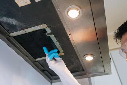 Person wearing protective gloves and safety glasses removes greasy stainless steel range hood filter for cleaning, emphasizing routine maintenance and home hygiene.
