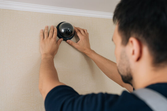 Over shoulder view of male technician installing black dome security camera. Man works on home safety system, mounting surveillance equipment on wall for protection and monitoring. Home improvement.