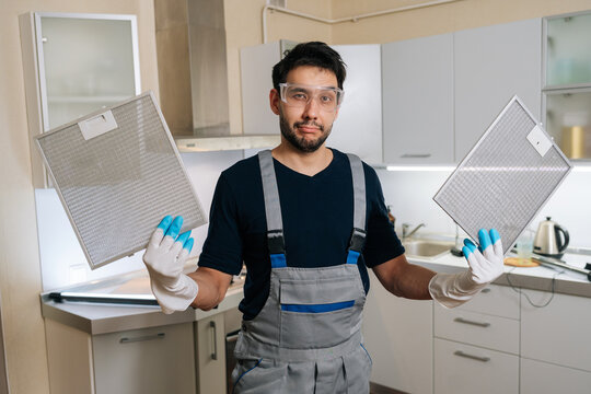 Confused male technician in uniform and goggles holding two dirty ventilation filters. Puzzled worker shrugs in modern kitchen. Concept for home repair, maintenance service cleaning.