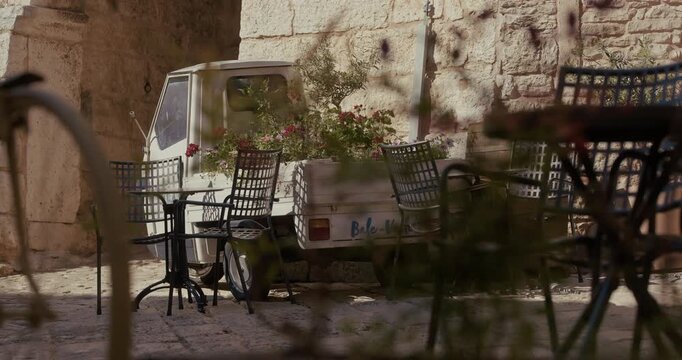 A small three-wheeled vehicle carrying flower pots is parked beside outdoor tables and chairs.