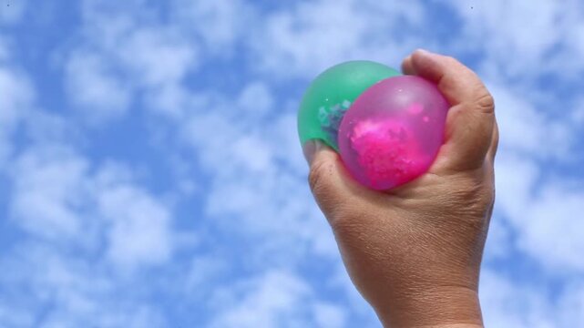 Close-up of one hand gripping and rotating transparent pink and green water-filled balloons against bright blue sky on sunny day, fingers pressing and releasing the elastic surface repeatedly.