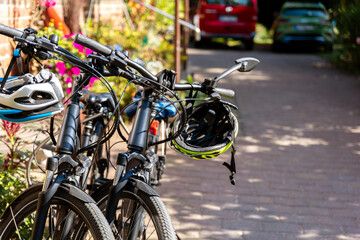 Fototapeta premium Bicycles stand parked sunny residential driveway helmets hanging handlebars garden flower suburban house. Scene eco friendly transport active lifestyle everyday cycling equipment calm outdoor setting