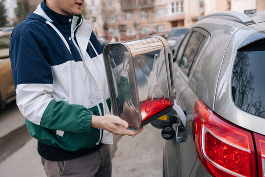 Man holding metal jerrycan, pouring gasoline into cars tank on city street, highlighting emergency refueling, fuel shortage and transportation challenges amid rising prices..