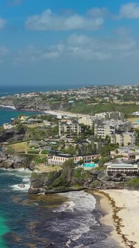 Vertical Screen: Stunning aerial view of a Caribbean coastal resort perched on rocky cliffs overlooking turquoise ocean waters and sandy beach with luxury buildings and palm trees.