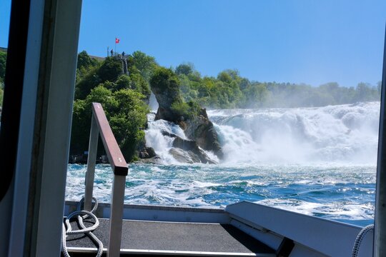 Rock formation in the middle of Rhine Falls with Swiss flag, surrounded by powerful flowing water and scenic natural landscape as seen from tourist boat