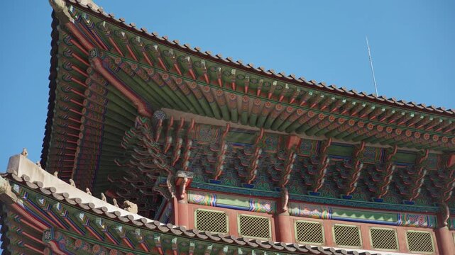 Stunning Gyeongbokgung Palace architecture featuring elaborate dancheong painted wooden brackets and eaves under clear blue skies, with golden Korean characters on signage and intricate roof details.
